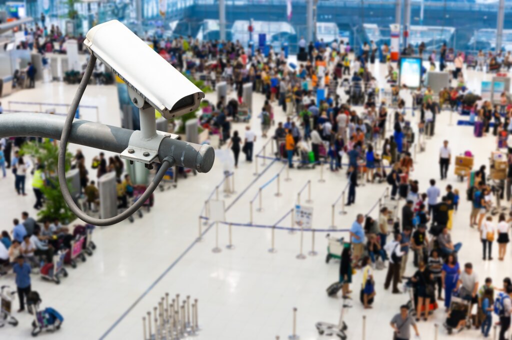 Busy airport waiting area with a CCTV camera in the foreground.